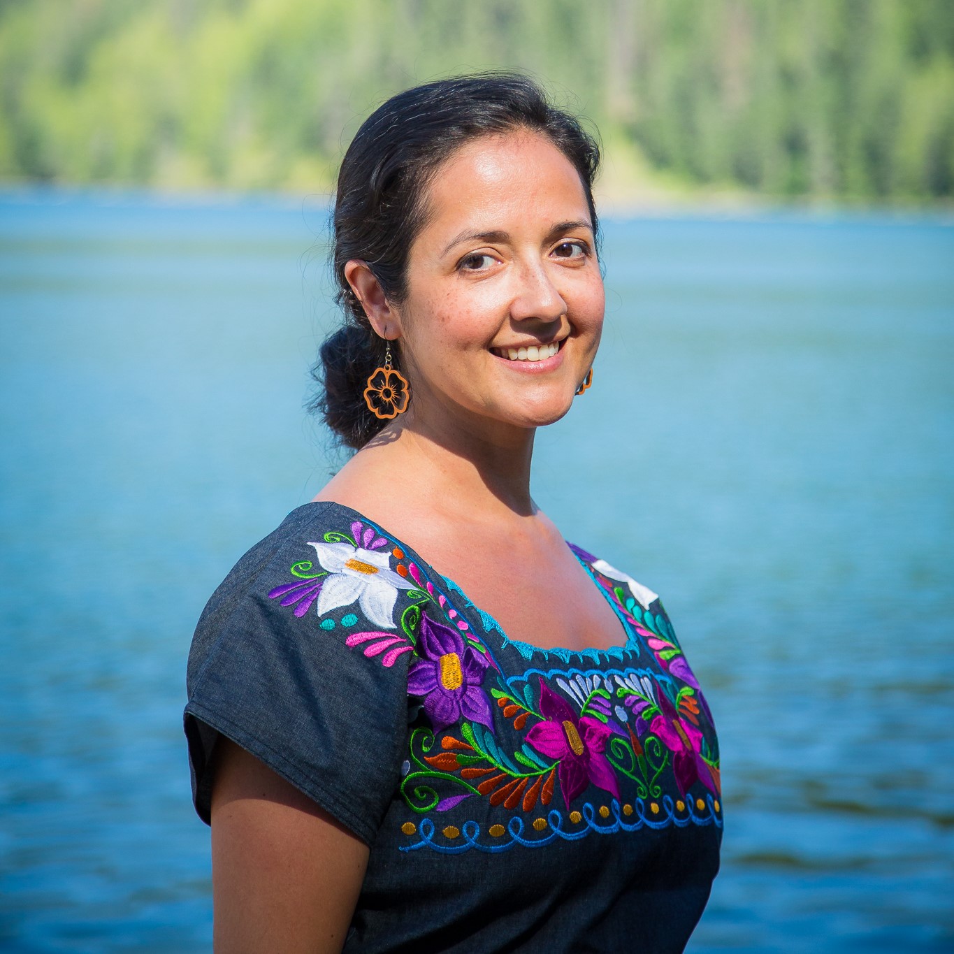 Clara is standing in front of a calm, blue lake that is lined by evergreen covered mountain tops on the not too distant shore. She is smiling warmly at the camera, and her dark brown hair is pulled back in a low, loose bun. She is wearing flower shaped lazer-cut wood earings, and a denim dress that is embroidered along the shoulders and square neckline with a traditional Mexican design of vivid, multi-colored flower patterns.